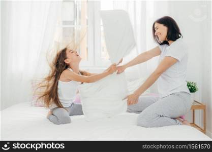 Parenthood and family concept. Attractive female mother with positive smile, spends free time with her daughter, have pillow fight, pose against bedroom interior, enjoys leisure time during weekend