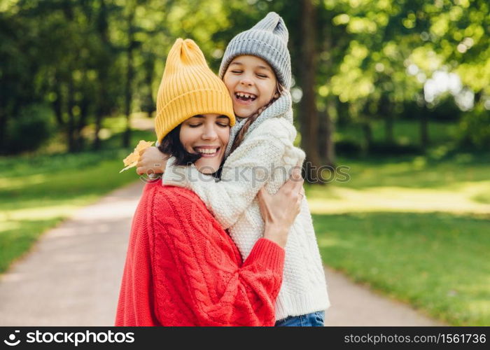 Parenthood, happiness and lifestyle concept. Beautiful female and her daughter have warm embrace, dressed in knitted clothes, have walk during autumn day, enjoy sunny weather, love each other