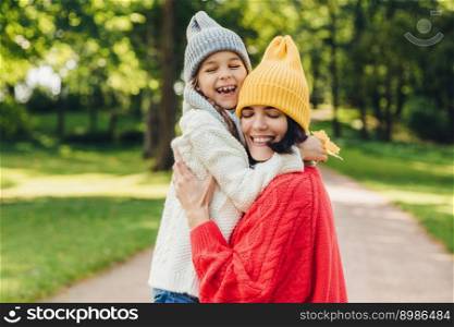Parenthood, happiness and lifestyle concept. Beautiful female and her daughter have warm embrace, dressed in knitted clothes, have walk during autumn day, enjoy sunny weather, love each other