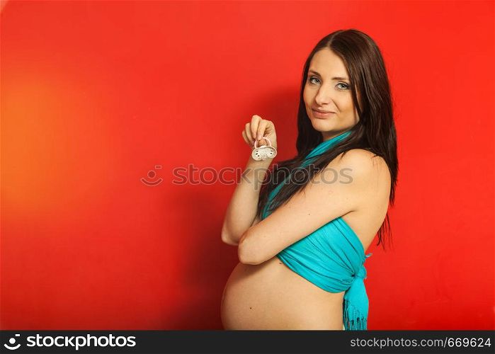 Parenthood, waiting for baby concept. Adult woman showing her big pregnant belly holding pacifier. Indoor shot on red background. Adult woman showing her pregnant belly