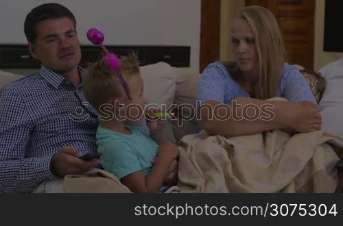 Parents and child at home. Mother and father trying to watch TV while child in funny ears playing with party horn and talking to mom