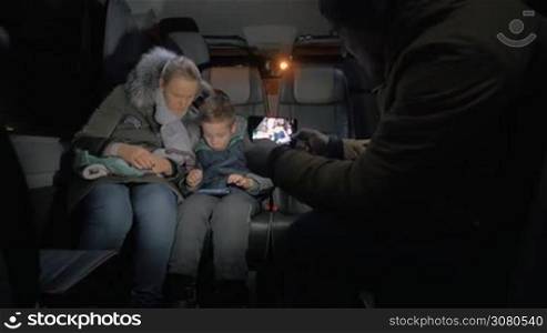 Parents and child traveling by minibus. Man shooting mobile video of mom and son playing game on smart phone