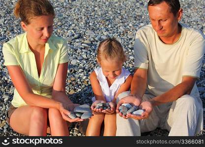 Parents and daughter sits in pebbly beach ind holding pebbles in hands