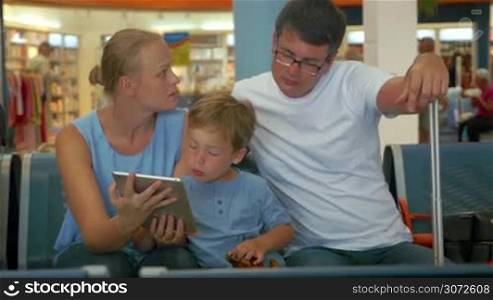 Parents and son are sitting in waiting room before departure. Son is searching something in tablet PC.