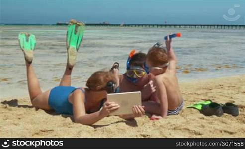 Parents and their son are lying on the beach after snorkel diving. They are watching something in tablet PC and talking.