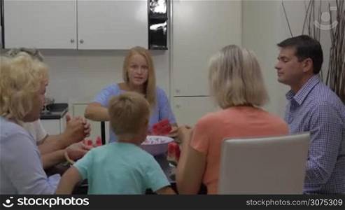Parents, grandparents and child eating watermelon in the kitchen. Adults sitting at the table, while child is running to mother to get a tasty fruit