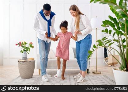Parents help a little girl with a broken leg, living room interior on background. Mother, father and their little daughter cope with the trouble together, parental care. Parents help a little girl with broken leg