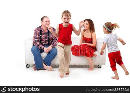 Parents sit on white leather sofa and look at running children
