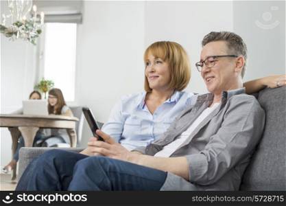 Parents watching TV on sofa with children using laptop in background