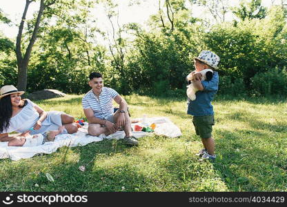 Parents with baby and toddler sons hugging soft toy on picnic blanket in Pelham Bay Park, Bronx, New York, USA