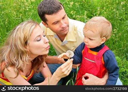 Parents with child sit in grass and blow on dandelion