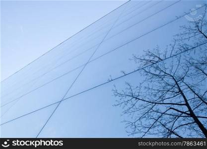 part of geometrically shaped modern glass building with reflections of blue sky and winter tree