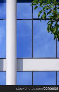 Part of white concrete column in front of blue glass wall with blurred green tree branch on foreground in vertical frame