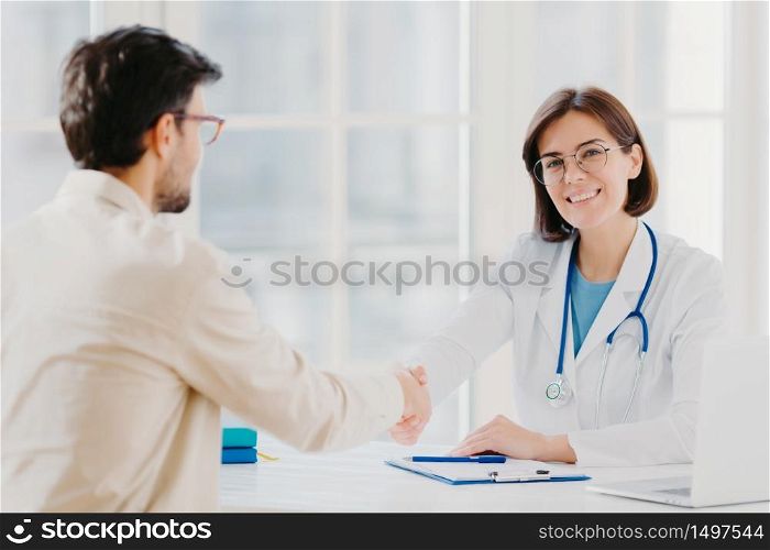 Partnership, assistance, trust and medicine concept. Female doctor shakes hands with thankful patient for good treatment and professionalism, pose in clinic, medical records near on white table