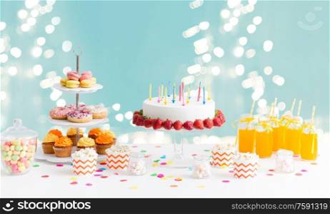party and festive concept - birthday cake with candles and strawberries, drinks and food on table over lights on blue background. food and drinks on table at birthday party