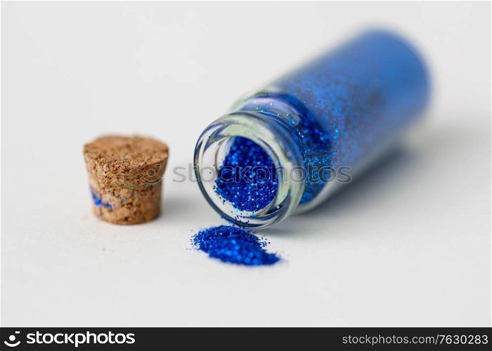 party, decoration and holidays concept - close up of ultramarine blue glitters poured from small glass bottle and cork stopper over white background. blue glitters poured from small glass bottle