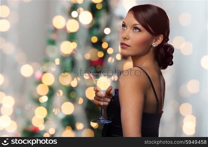 party, drinks, holidays, luxury and celebration concept - woman in evening dress with cocktail over christmas tree lights