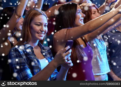 party, holidays, celebration, nightlife and people concept - smiling young woman with smartphone texting message at concert in club and snow effect