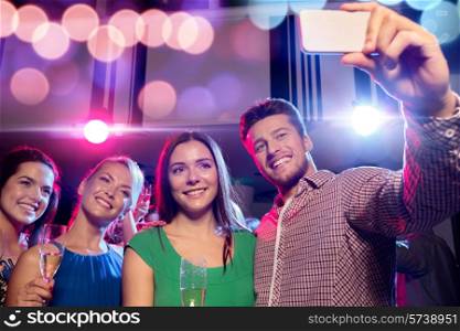 party, holidays, technology, nightlife and people concept - smiling friends with glasses of champagne and smartphone taking selfie in club