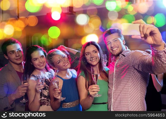 party, holidays, technology, nightlife and people concept - smiling friends with glasses of non-alcoholic champagne and smartphone taking selfie in club