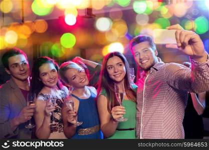 party, holidays, technology, nightlife and people concept - smiling friends with glasses of non-alcoholic champagne and smartphone taking selfie in club