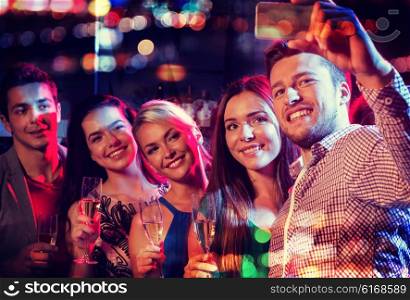 party, holidays, technology, nightlife and people concept - smiling friends with glasses of champagne and smartphone taking selfie in night club with holidays lights