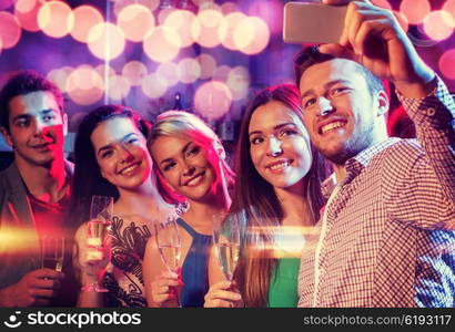 party, holidays, technology, nightlife and people concept - smiling friends with glasses of champagne and smartphone taking selfie in club