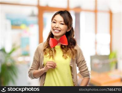 party props and people concept - happy asian young woman with big red bow tie over office background. happy asian woman with big red bow tie