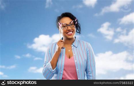 party props, photo booth and people concept - happy african american young woman with big glasses over blue sky and clouds background. happy african american woman with big glasses