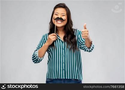 party props, photo booth and people concept - happy asian young woman with big black moustaches showing thumbs up over grey background. asian woman with vintage moustaches party prop