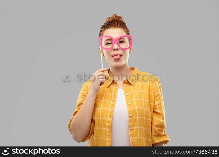 party props, photo booth and people concept - smiling red haired teenage girl in checkered shirt with big pink glasses sticking her tongue out over grey background. smiling red haired teenage girl with big glasses
