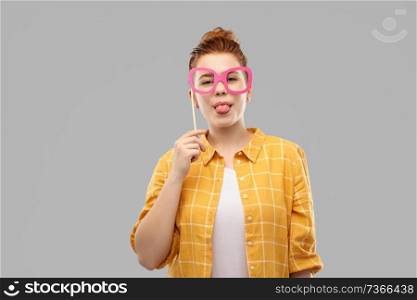 party props, photo booth and people concept - smiling red haired teenage girl in checkered shirt with big pink glasses sticking her tongue out over grey background. smiling red haired teenage girl with big glasses