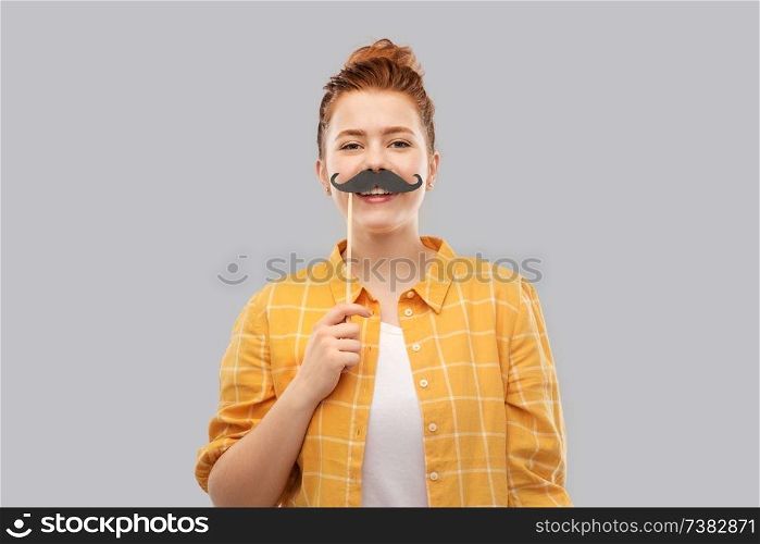 party props, photo booth and people concept - smiling red haired teenage girl in checkered shirt with big black moustaches over grey background. red haired teenage girl with black moustaches