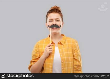 party props, photo booth and people concept - smiling red haired teenage girl in checkered shirt with big black moustaches over grey background. red haired teenage girl with black moustaches