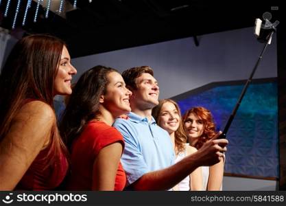 party, technology, nightlife and people concept - smiling friends with smartphone and monopod taking selfie in club