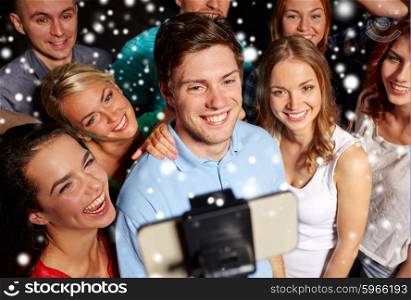 party, technology, nightlife and people concept - smiling friends with smartphone and monopod taking selfie in club and snow effect