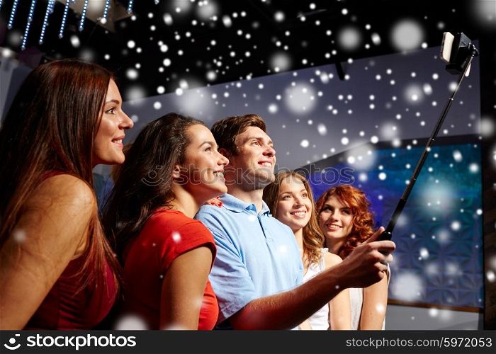 party, technology, nightlife and people concept - smiling friends with smartphone and monopod taking selfie in club and snow effect
