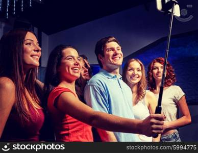 party, technology, nightlife and people concept - smiling friends with smartphone and monopod taking selfie in club