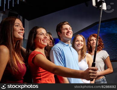 party, technology, nightlife and people concept - smiling friends with smartphone and monopod taking selfie in club