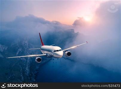 Passenger airplane is flying over clouds at sunset. Landscape with white airplane, low clouds, sea coast, purple sky at dusk. Aircraft is landing. Business trip. Commercial plane. Travel. Aerial view