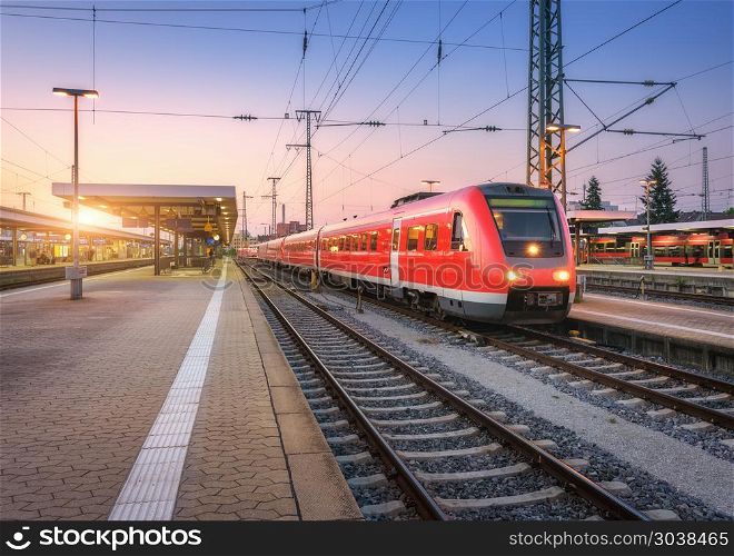 Passenger high speed train on the railway station at sunset. Urban landscape with modern commuter train on the railway platform with illumination at dusk. Intercity vehicle on railroad in Europe. Passenger high speed train on the railway station at sunset