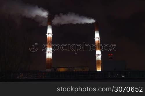 Passenger train in the city at night. It passing by factory with two smoking pipes