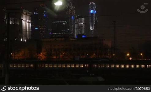 Passenger train passing slowly through the city at night. Modern illuminated skyscrapers in background