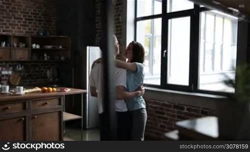 Passionate couple hugging, looking at each other with love while dancing together in slow dance in modern kitchen in the morning. Young couple in love sharing the beautiful moments of togetherness dancing. Slow motion. Steadicam stabilized shot.