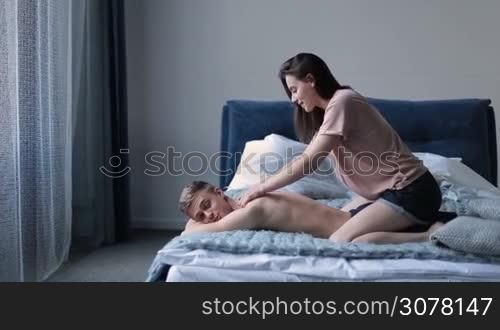 Passionate woman doing massage to her handsome boyfriend lying on bed at home. Attractive couple enjoying nice morning together. Beautiful brunette making massage to happy young man in bedroom. Side view.