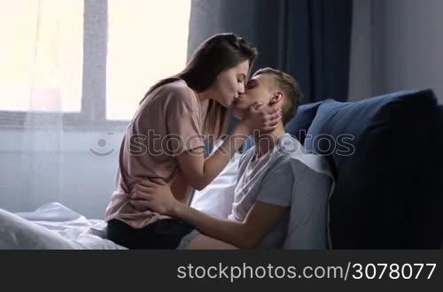 Passionate young attractive couple embracing and kissing while relaxing in bed after waking up in the morning. Romantic couple in bed being intimate. Side view.