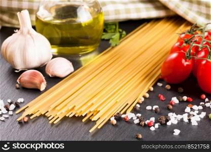 Pasta and cooking ingridients on black slate surface. Spaghetti, tomato, olive oil, garlic, parsley and spices. Italian cuisine. Pasta and food ingredients on black background.
