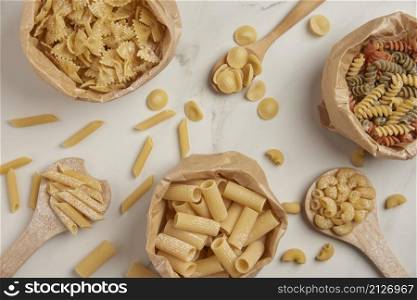 pasta concept some types of pasta being organized on wooden spoons and in paper bags while some and other ingredients being put on white background.