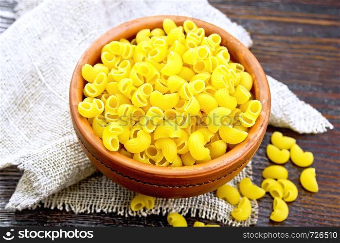 Pasta Elbow macaroni in a clay bowl on napkin of burlap on the background of dark wooden board