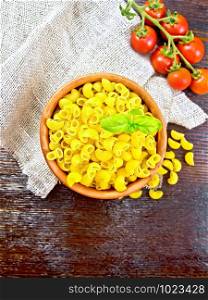 Pasta Elbow with basil in a clay bowl on napkin of burlap, tomatoes on wooden board background from above